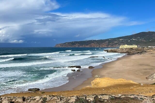 Waves crashing against the rocky shore at Praia do Guincho with Fortaleza do Guincho in the background